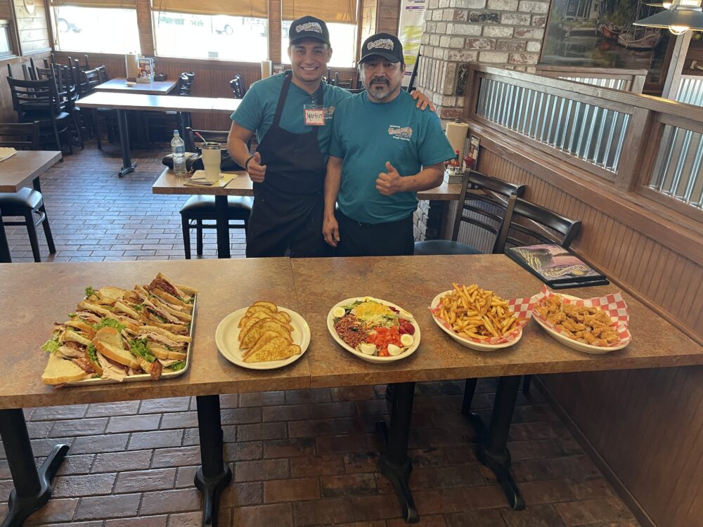 Two men standing behind a table full of different plates of food.